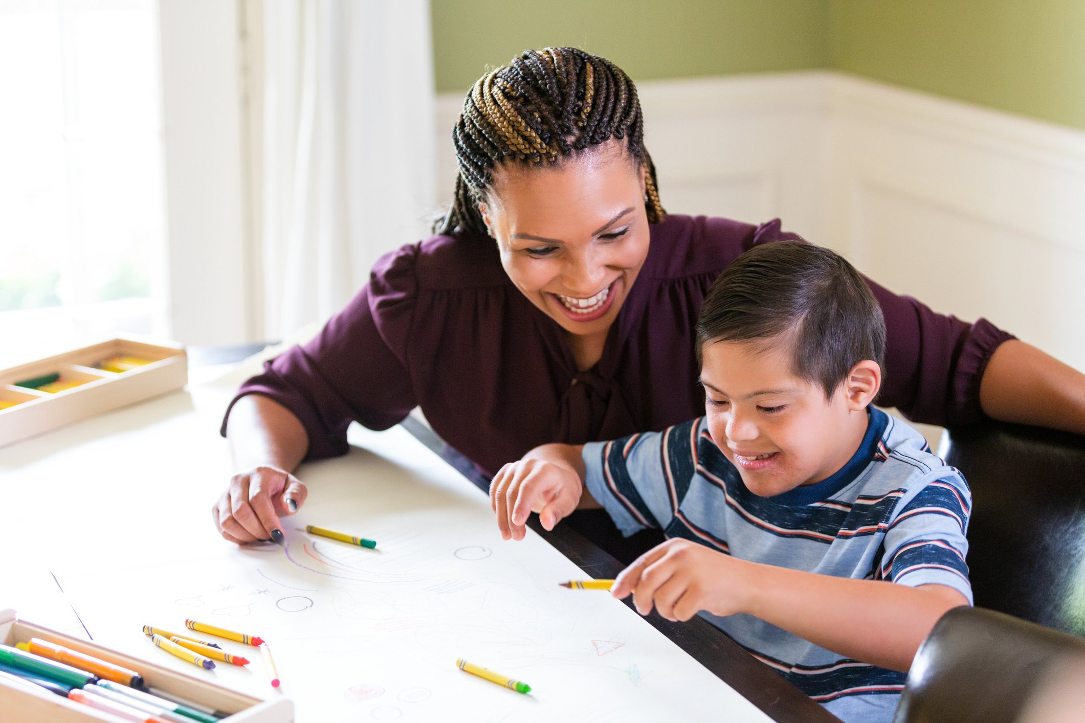 woman smiling watching a young boy with disabilities draw with crayons